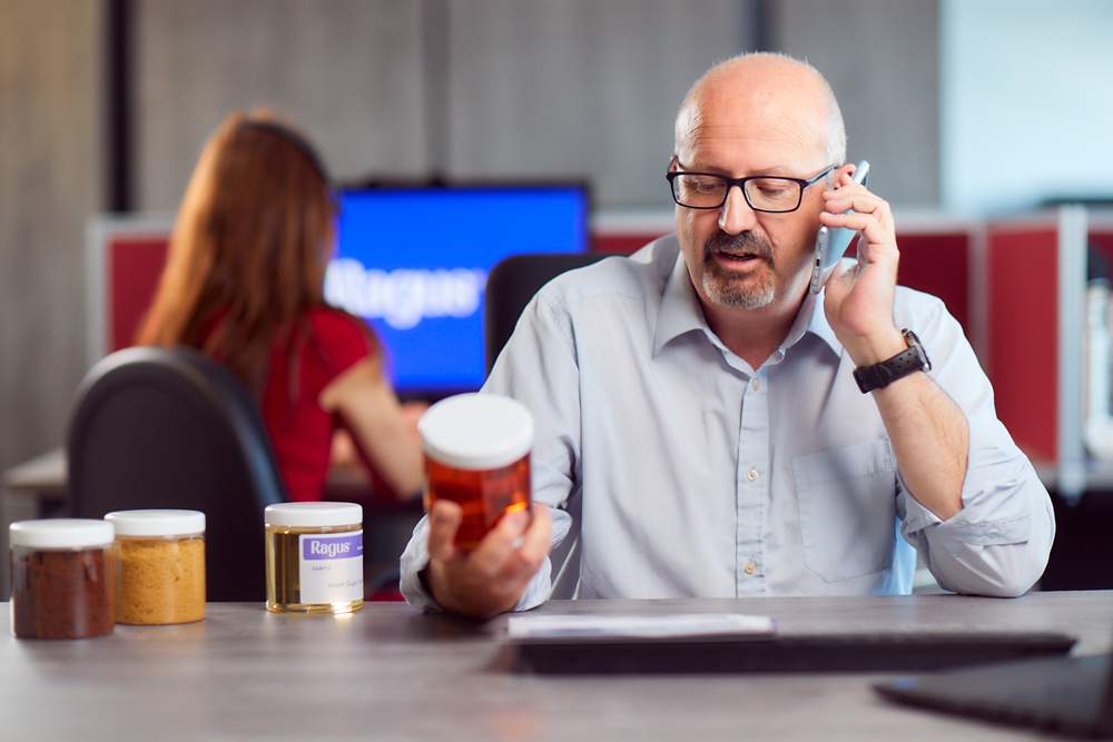A man in an office setting sat behind a desk on the phone while looking at sugar products in glass jars