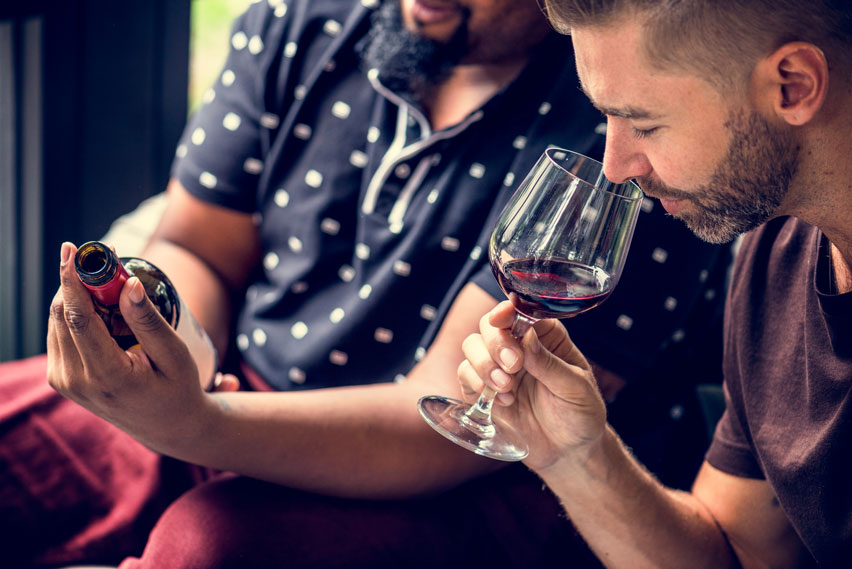 Two men sat next to each other, one is holding a wine bottle and the other is sniffing a small glass of read wine