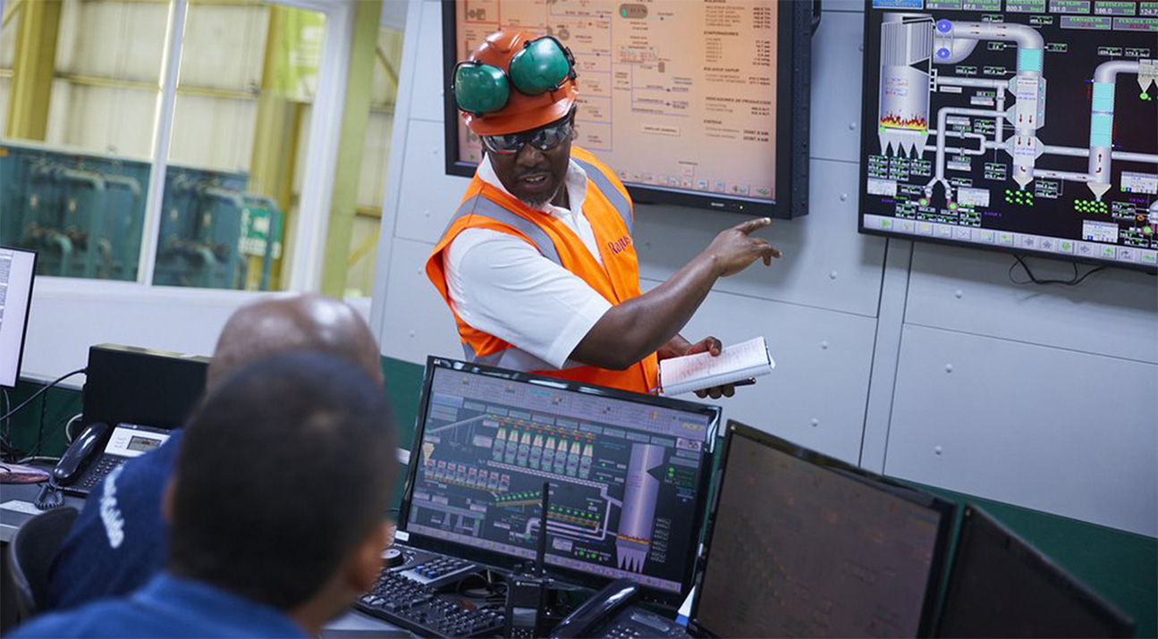 A man in an orange high visibility jacket stands at the front of a control room pointing at a screen