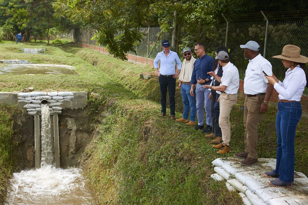 A group of men stand in front of a small stream running through a plastic pipe into a ditch